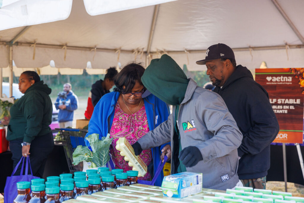 A Goodr team member in a gray jacket helps a woman place eggs in her shopping bag with her collard greens and other groceries