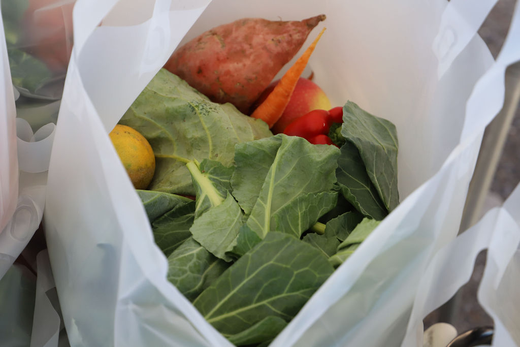A plastic produce bag filled with collard greens, sweet potatoes, lemons, and other fresh produce