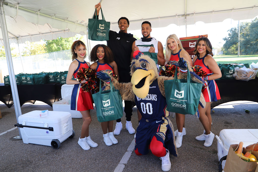 Pelicans players CJ McCollum and Trey Murphy III, Pierre the Pelican, and Pelican dancers holding green bags filled with shelf-stable groceries at CJ McCollum's McCollum Friendsgiving Pop-Up Grocery Market 2023